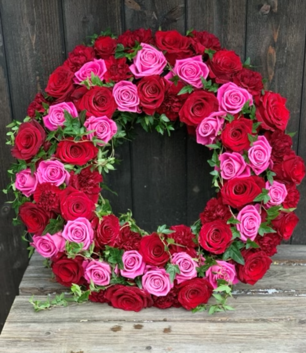 Rundbunden krans med vackra rosa, röda rosor, röd nejlika och murgröna. Till denna rekomenderar vi ett fint ljusrosa band alt silvergrönt eller rött.
